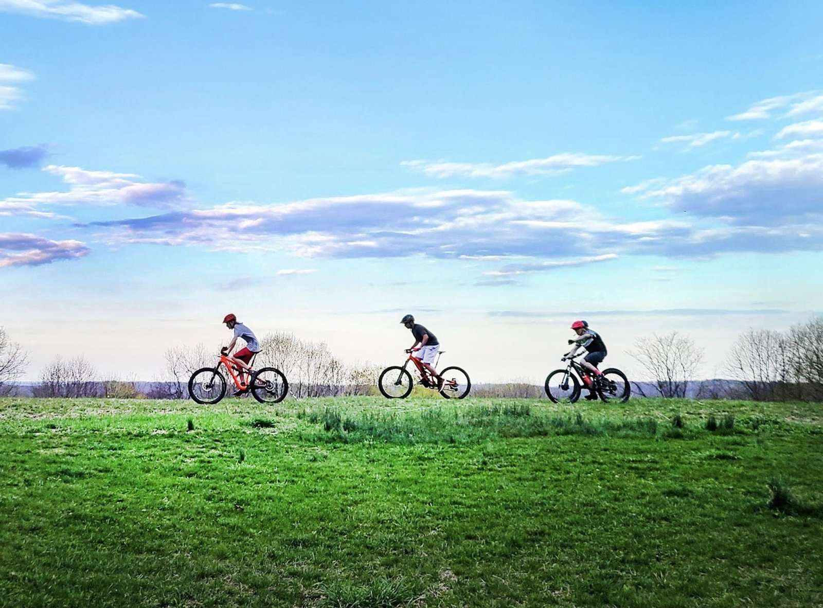 Three cyclists riding mountain bikes across a green hilltop under blue sky